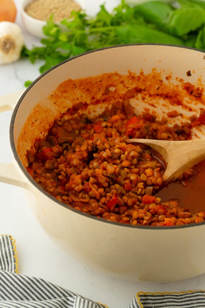 A beige pot filled with cooked lentil stew containing diced vegetables, stirred with a wooden spoon. Fresh herbs, garlic, and an onion are visible in the background—ingredients often associated with habits for longevity.