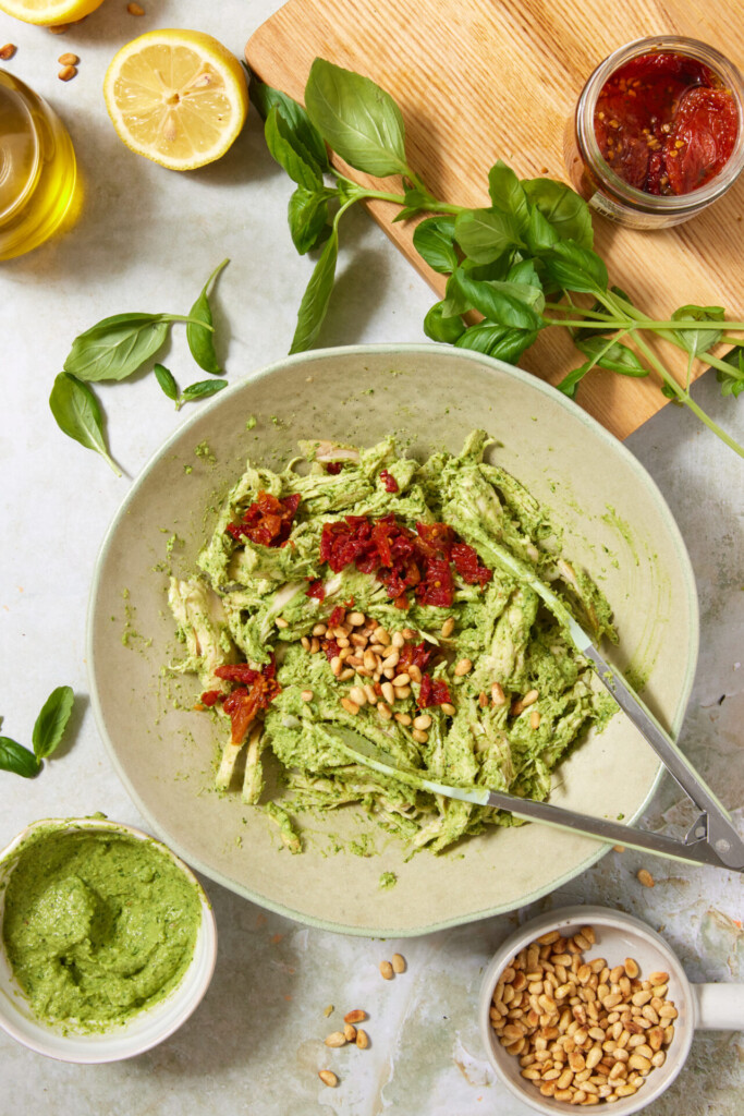 A bowl of pasta with green pesto sauce, sun-dried tomatoes, and pine nuts, surrounded by fresh basil, pine nuts, half a lemon, pesto in a bowl, olive oil, and a wooden cutting board.