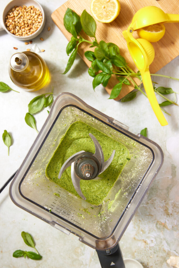A blender with green pesto, surrounded by fresh basil, olive oil, pine nuts, a lemon half, a yellow lemon squeezer, and scattered basil leaves on a light surface.