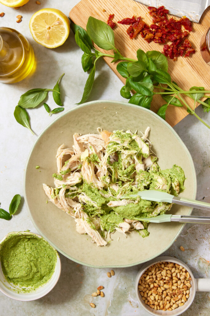 A bowl of shredded chicken mixed with green pesto, surrounded by fresh basil, pine nuts, sun-dried tomatoes, olive oil, a lemon half, and a small bowl of pesto on a light countertop.