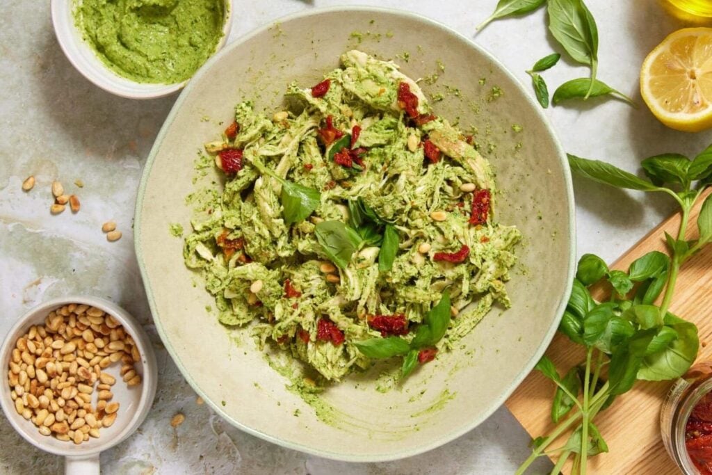 A bowl of pasta with green pesto, sun-dried tomatoes, and basil. Surrounding items include lemons, basil leaves, pine nuts, olive oil, pesto sauce, and a wooden board with sun-dried tomatoes in a jar.