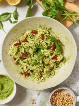A bowl of pasta with green pesto, sun-dried tomatoes, and basil. Surrounding items include lemons, basil leaves, pine nuts, olive oil, pesto sauce, and a wooden board with sun-dried tomatoes in a jar.