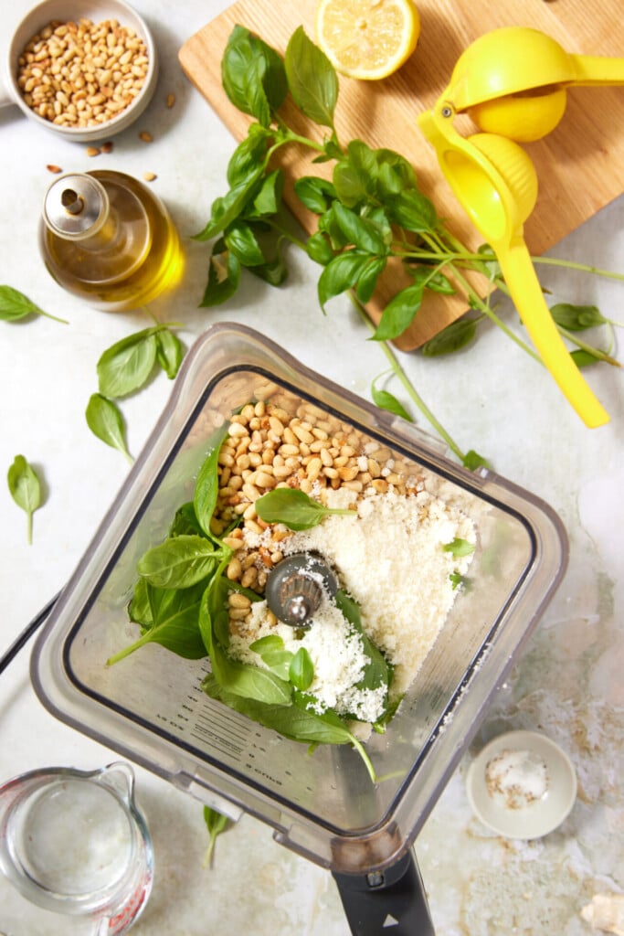 Overhead view of a blender with basil leaves, pine nuts, and grated Parmesan. Surrounding items include fresh basil, lemon, olive oil, water, pine nuts, and a lemon squeezer on a light kitchen counter.