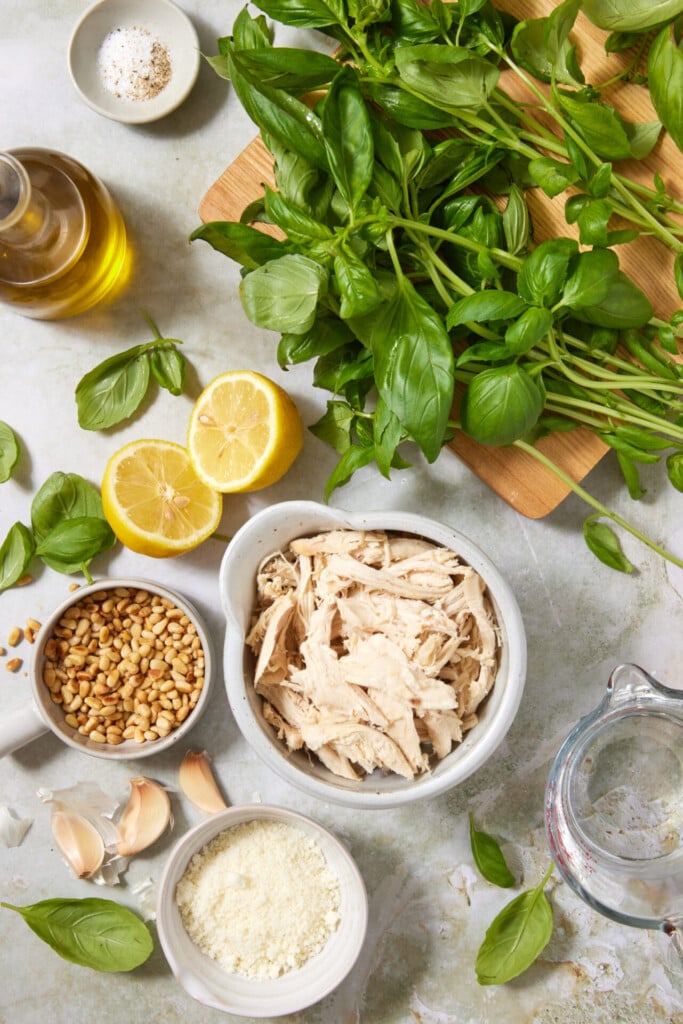 A flatlay of shredded chicken, fresh basil, lemon halves, pine nuts, grated cheese, garlic cloves, olive oil, salt, and water on a light surface, with some basil on a cutting board.