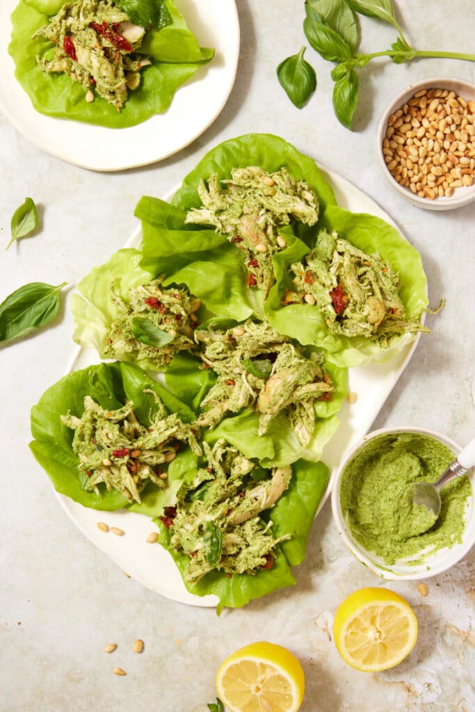 A plate of lettuce wraps filled with shredded chicken and green pesto, garnished with sun-dried tomatoes and pine nuts. Fresh basil, lemon halves, pine nuts, and a bowl of pesto are nearby on the table.