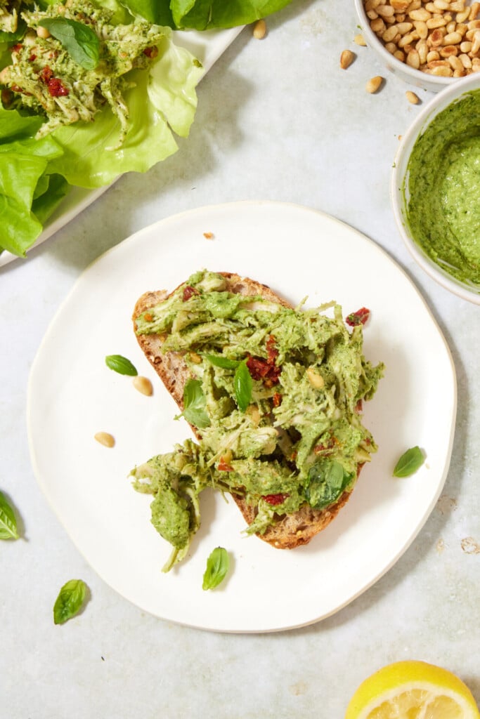 Slice of bread topped with shredded chicken mixed with green pesto, basil leaves, pine nuts, and sun-dried tomatoes on a white plate. Surrounding items include a bowl of pesto, pine nuts, lettuce, and a lemon slice.