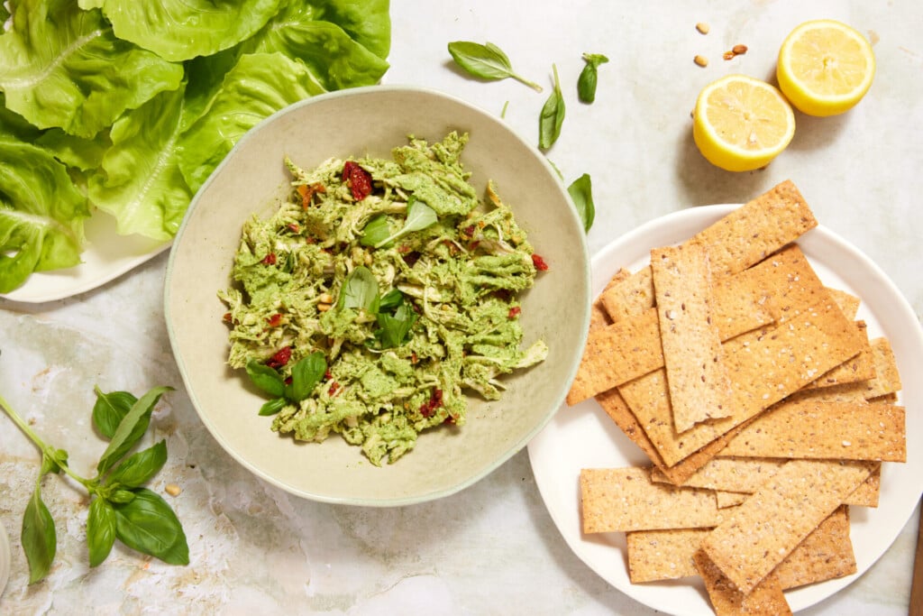 A bowl of green pesto chicken salad with sun-dried tomatoes, fresh basil, lettuce leaves, lemon halves, and a plate of crispbread crackers on a light surface.