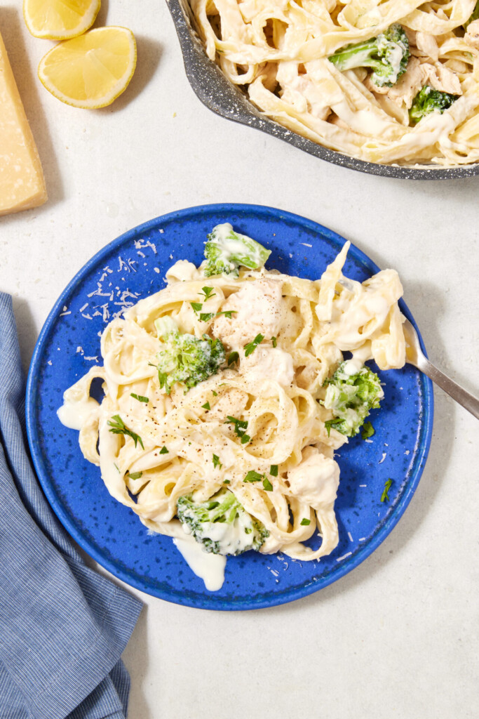 A blue plate with high-protein Cottage Cheese Fettuccine Alfredo, broccoli, and chicken, garnished with black pepper and herbs. A fork rests on the plate. Nearby are lemon wedges, Parmesan cheese, and a blue napkin.