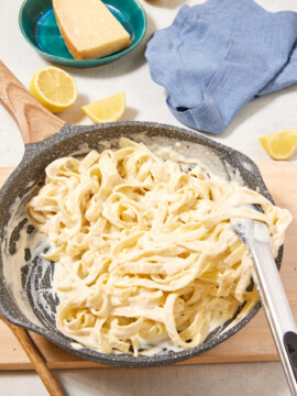 A pan of creamy high-protein Cottage Cheese Fettuccine Alfredo sits on a wooden board with metal tongs. Lemon wedges, a block of cheese on a plate, a blue cloth, and a wooden spoon are nearby.