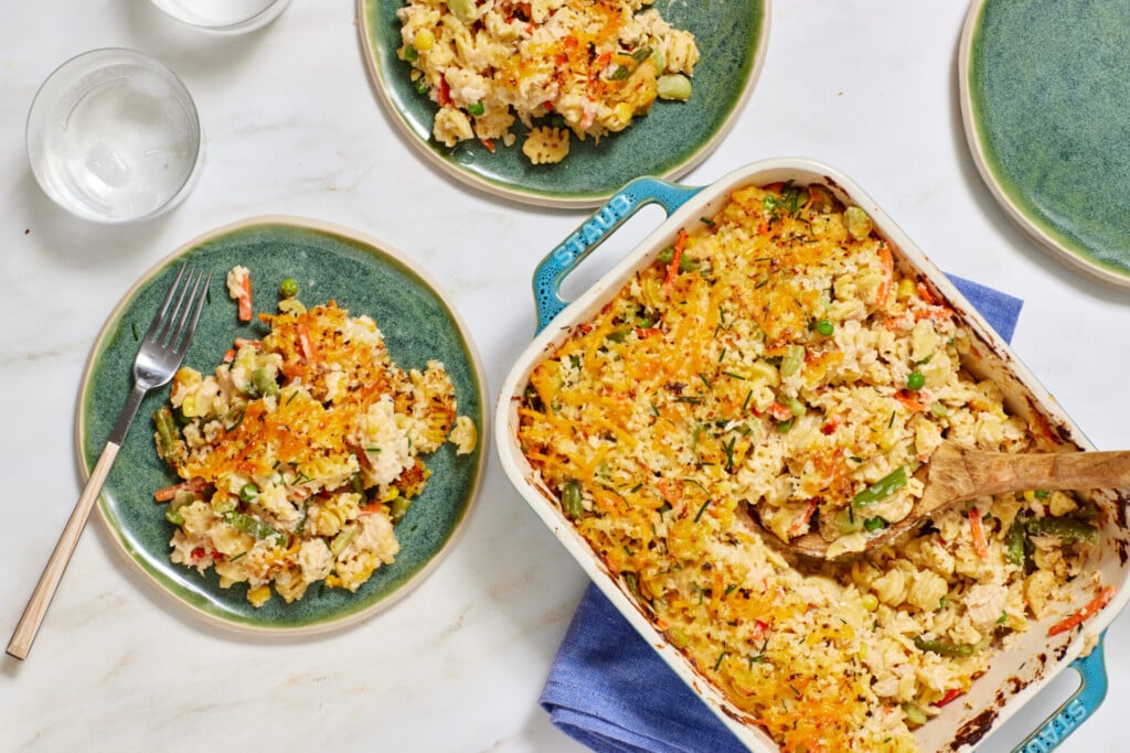 A casserole dish filled with baked pasta, vegetables, and cheese sits on a table. Three plates hold servings of the casserole, and there are two glasses of water nearby. A wooden spoon rests in the casserole dish.