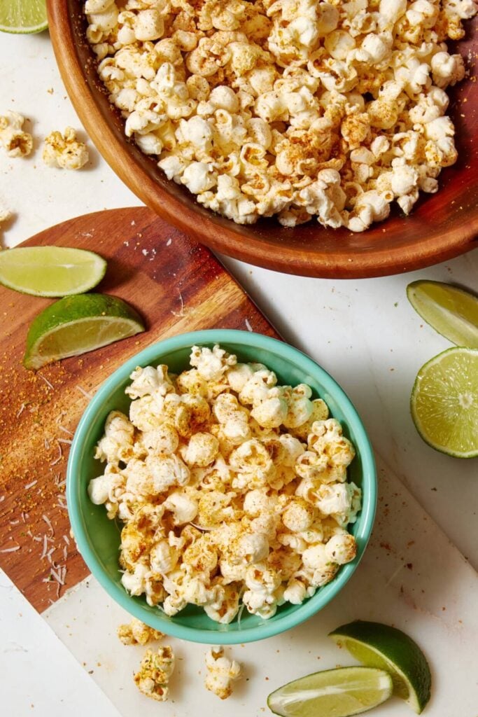 Two bowls of Fiesta Lime Popcorn sit on a white surface, surrounded by lime wedges and a wooden board dusted with seasoning.