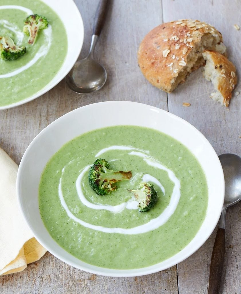 Two bowls of healthy broccoli soup garnished with a swirl of cream and roasted broccoli florets, served with a sliced bread roll.