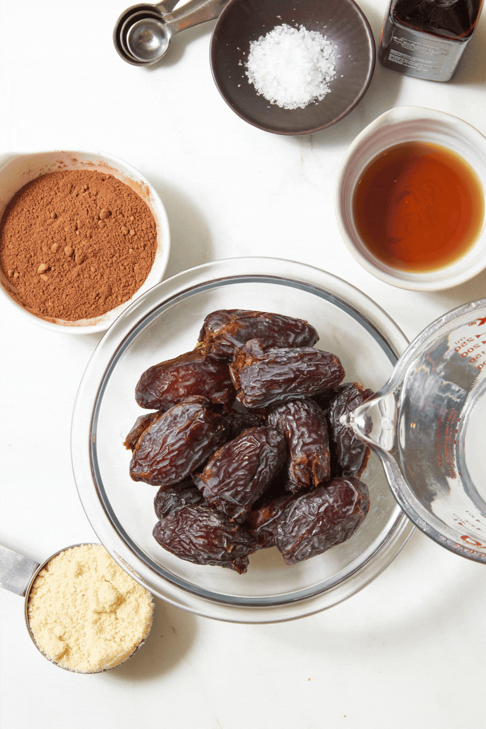 A glass bowl of whole dates for chewy chocolate caramel candies surrounded by small bowls of cocoa powder, salt, maple syrup, almond flour, and a measuring cup of water.