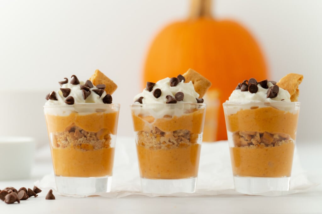 Three shot glasses filled with layers of healthy fall recipes, featuring pumpkin mousse and crushed graham crackers, topped with whipped cream, chocolate chips, and a piece of graham cracker, with a pumpkin in the background.