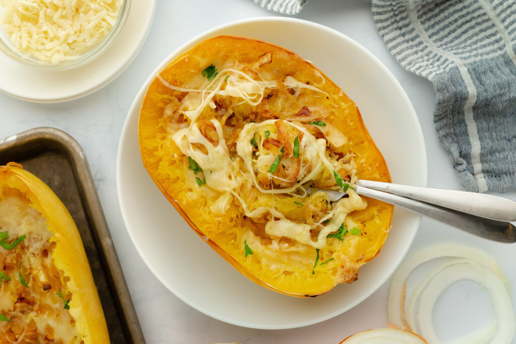 A French onion spaghetti squash half topped with melted cheese and herbs in a white bowl, with a fork pulling at the strands.