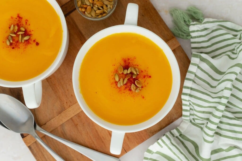 Two bowls of healthy pumpkin soup topped with seeds and paprika, served on a wooden board with spoons and a striped napkin.