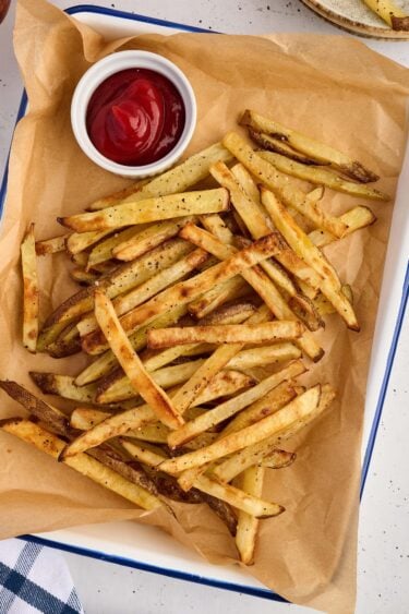 A pile of oven baked French fries, thin-cut and seasoned with pepper on parchment paper, served with a small bowl of ketchup.
