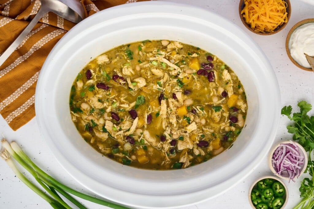 A white slow cooker filled with chicken and beans in a savory broth, surrounded by bowls of toppings for healthy fall recipes including shredded cheese, sour cream, red onion, and jalapeños.