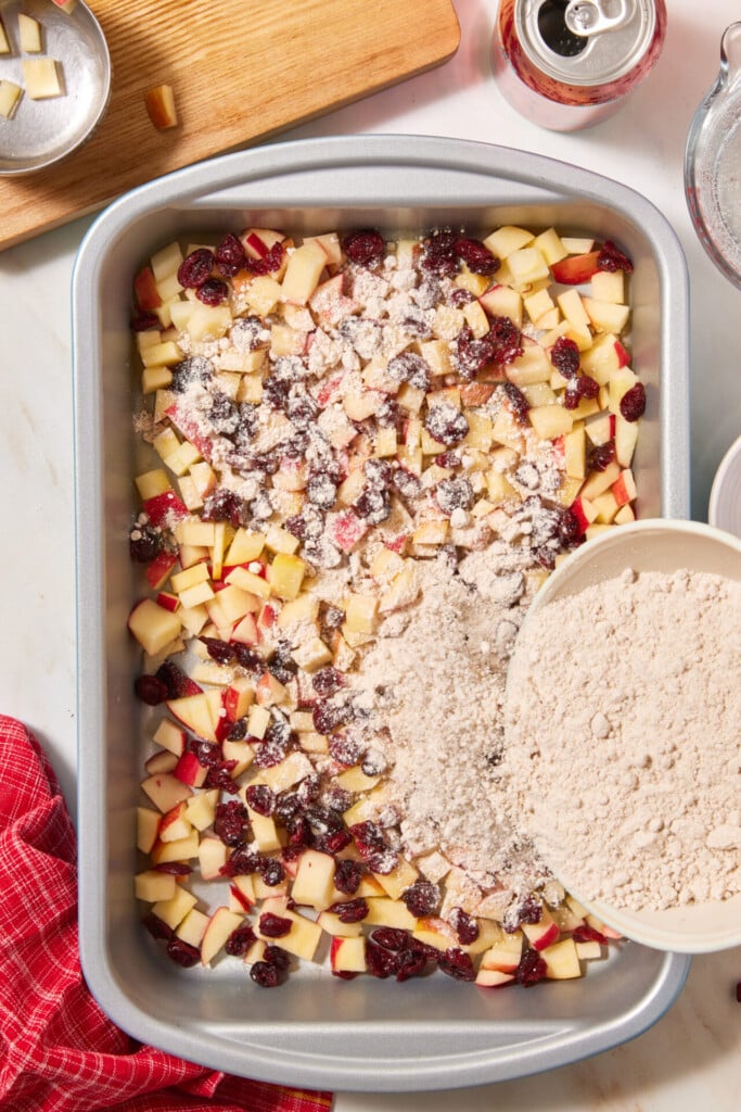 A bowl pouring dry crumble topping over chopped apples and dried cranberries in a metal baking pan for an easy apple cranberry dump cake.