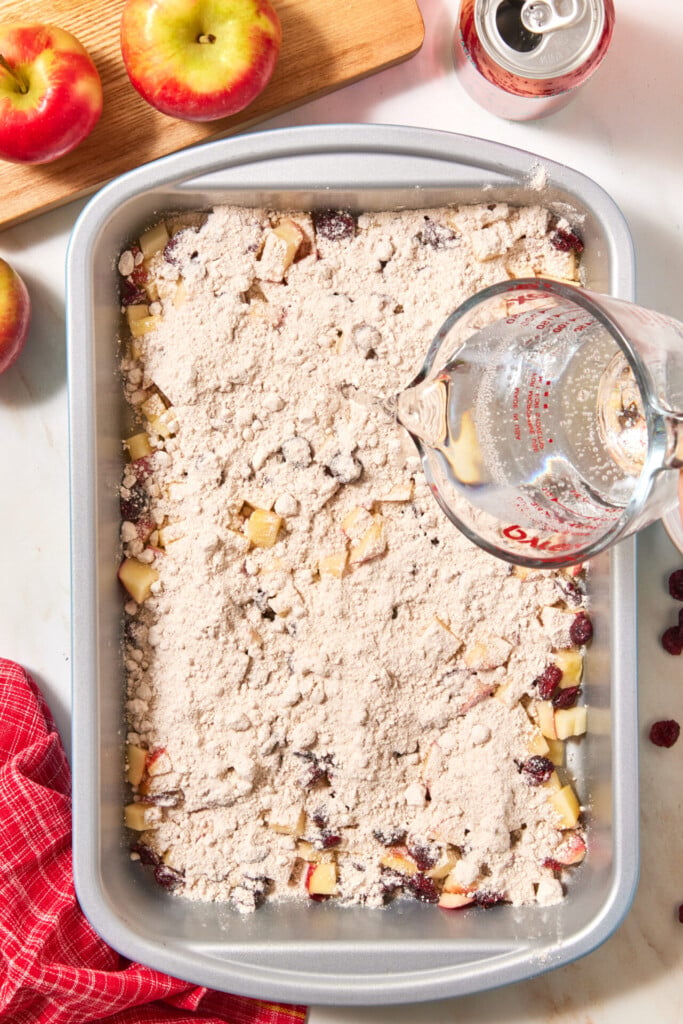 A glass measuring cup pours water over a baking pan filled with diced apples and dry crumble topping for an easy apple cranberry dump cake.