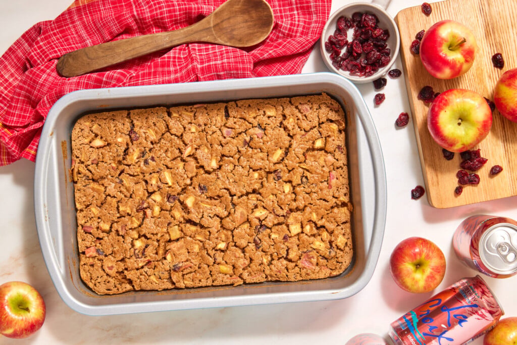 An easy apple cranberry dump cake sits in a rectangular metal baking pan, surrounded by whole apples, dried cranberries, a wooden spoon on a red cloth, and two cans of sparkling water.