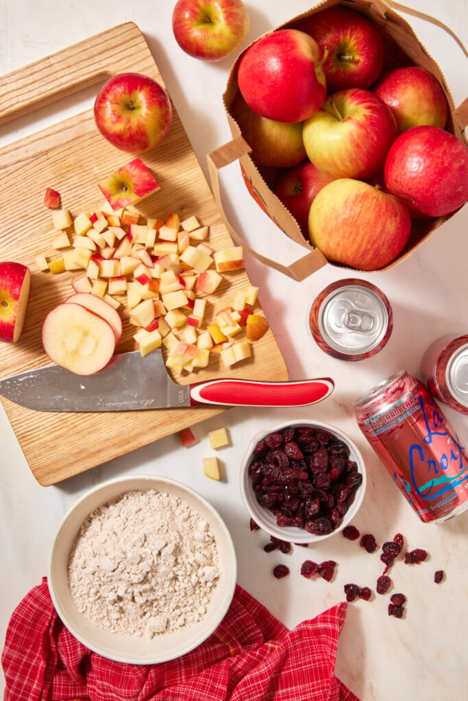 A wooden cutting board with a knife and diced apples for an easy apple cranberry dump cake, alongside a bag of whole apples, bowls of flour and dried cranberries, and a can of LaCroix sparkling water on a white surface.
