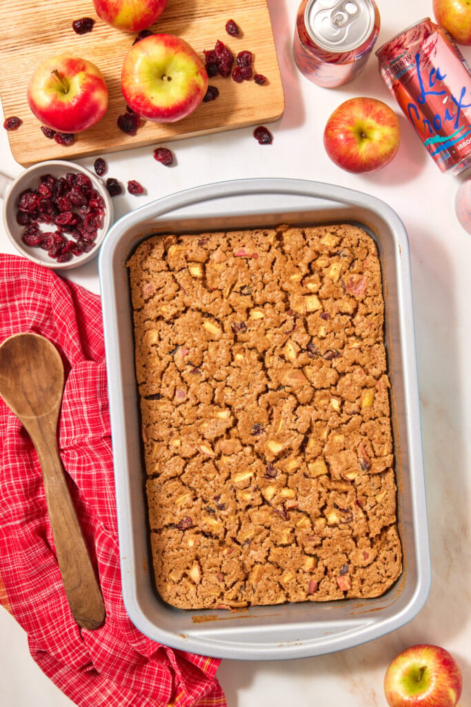A rectangular baking pan filled with an easy apple cranberry dump cake, surrounded by fresh apples, dried cranberries, a wooden spoon, and a can of sparkling water.