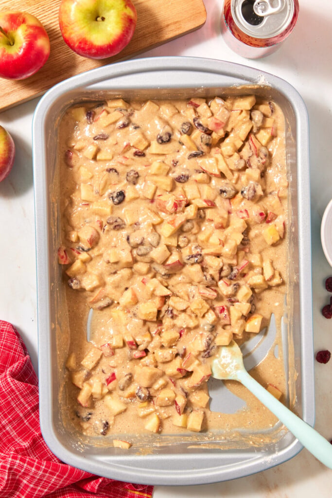 A rectangular metal baking pan filled with thick, raw apple cranberry dump cake batter containing chopped apples and dark raisins, with a silicone spatula resting inside.
