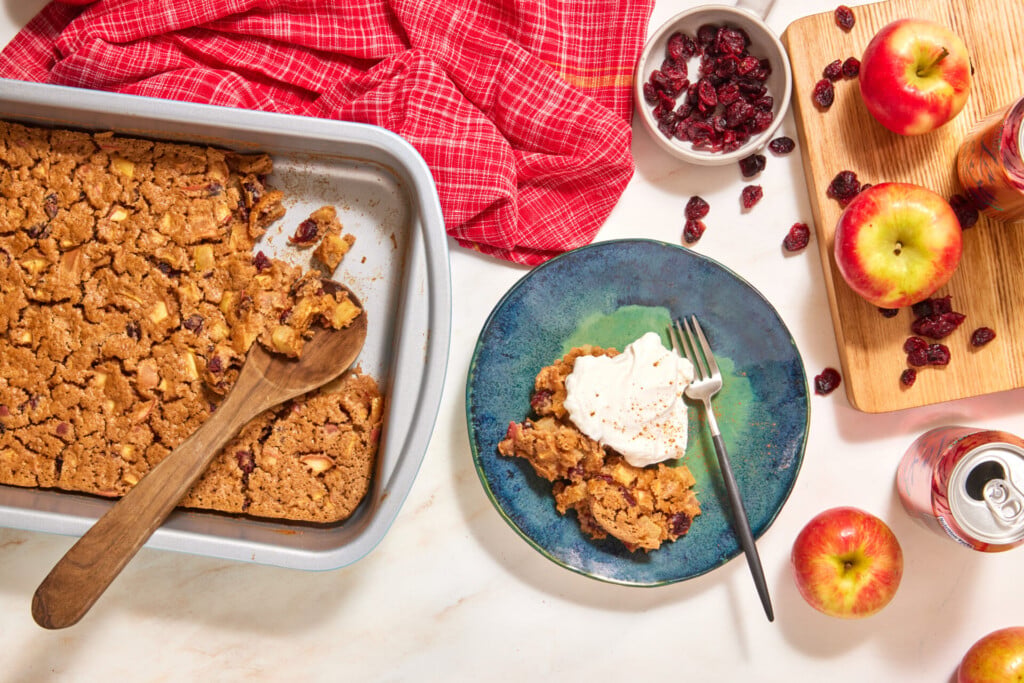 A metal baking pan containing an easy apple cranberry dump cake with a wooden spoon, and a serving topped with whipped cream on a blue plate, with whole apples and dried cranberries nearby.