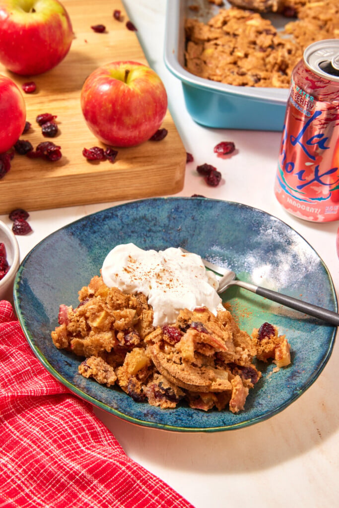 A serving of easy apple cranberry dump cake topped with whipped cream in a teal bowl, with fresh apples, a baking dish, and a can of sparkling water nearby.