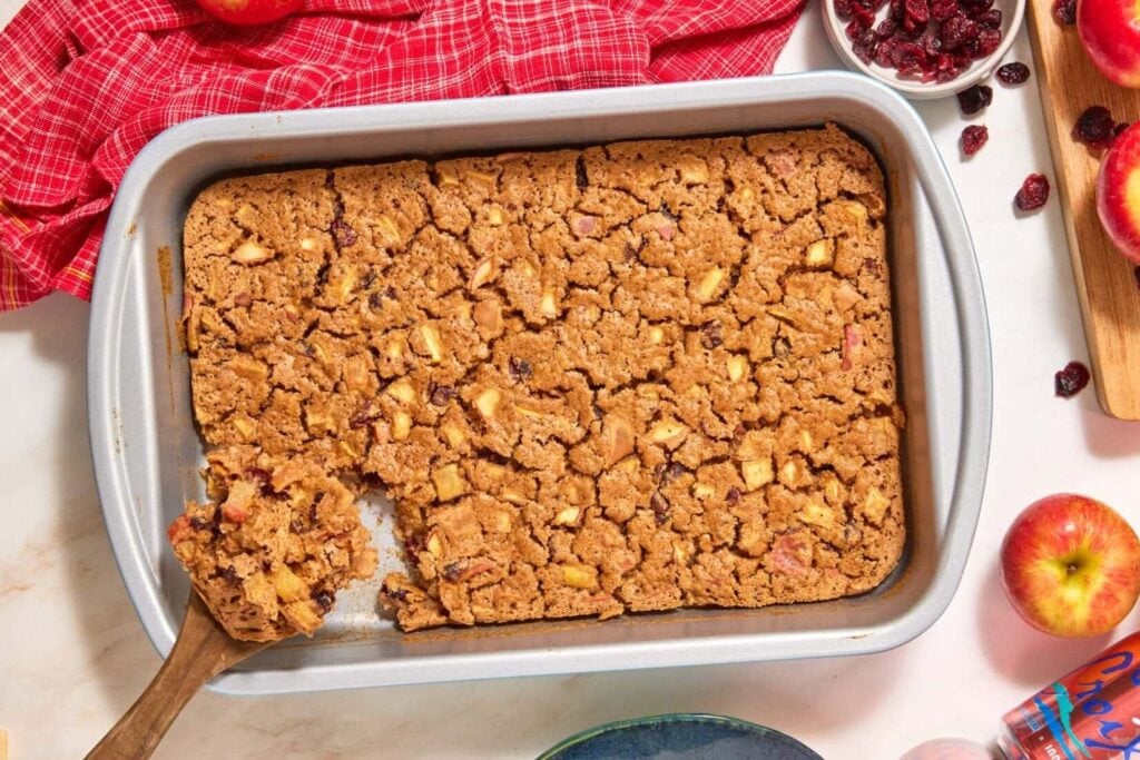 A rectangular pan of easy apple cranberry dump cake with a wooden spoon lifting a piece, surrounded by fresh apples, a small bowl of dried cranberries, and a can of sparkling water on a red plaid cloth.