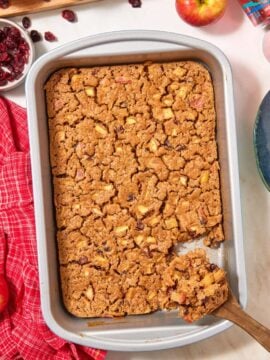 A rectangular pan of easy apple cranberry dump cake with a wooden spoon lifting a piece, surrounded by fresh apples, a small bowl of dried cranberries, and a can of sparkling water on a red plaid cloth.