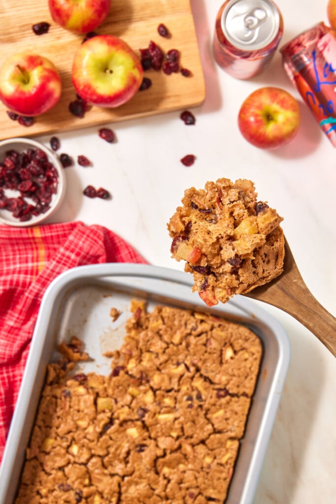 A wooden spoon lifts a portion of an easy apple cranberry dump cake above a square baking pan, surrounded by fresh apples and dried cranberries on a white surface.