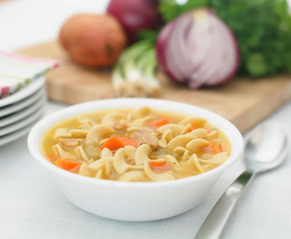 A white bowl of chicken noodle soup with carrots, next to a spoon and a cutting board with red onions and herbs, perfect for healthy soup recipes.