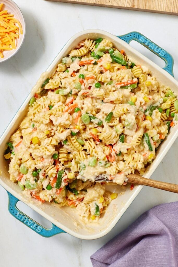 A casserole dish filled with creamy rotini pasta, chicken, and mixed vegetables. A wooden spoon rests inside. A bowl of shredded cheese and a pot with sauce are nearby on a white surface.