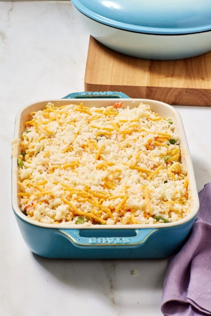 A blue casserole dish filled with an unbaked, cheesy vegetable casserole sits on a white countertop next to a matching blue pot and a folded lavender napkin.