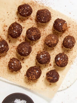 Fourteen chewy chocolate caramel candies made with dates, dusted with cocoa powder and topped with sea salt flakes, arranged on a sheet of parchment paper.