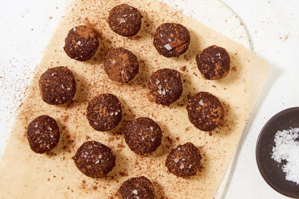 Fourteen chewy chocolate caramel candies made with dates, dusted with cocoa powder and topped with sea salt flakes, arranged on a sheet of parchment paper.