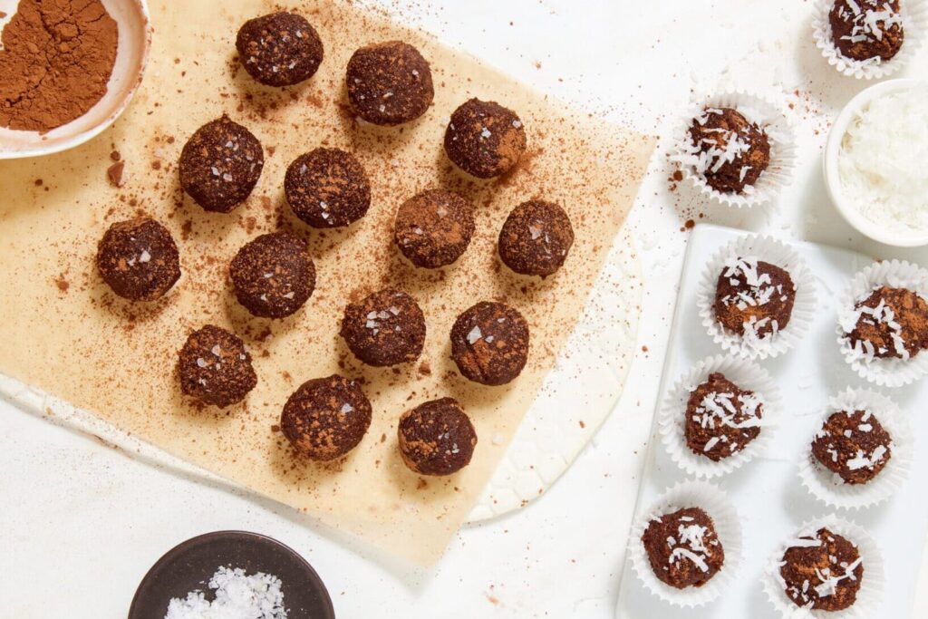 Overhead view of chewy chocolate caramel candies dusted with cocoa on parchment paper and coconut-topped chocolate balls in white paper liners, with bowls of cocoa powder, coconut flakes, and sea salt nearby.