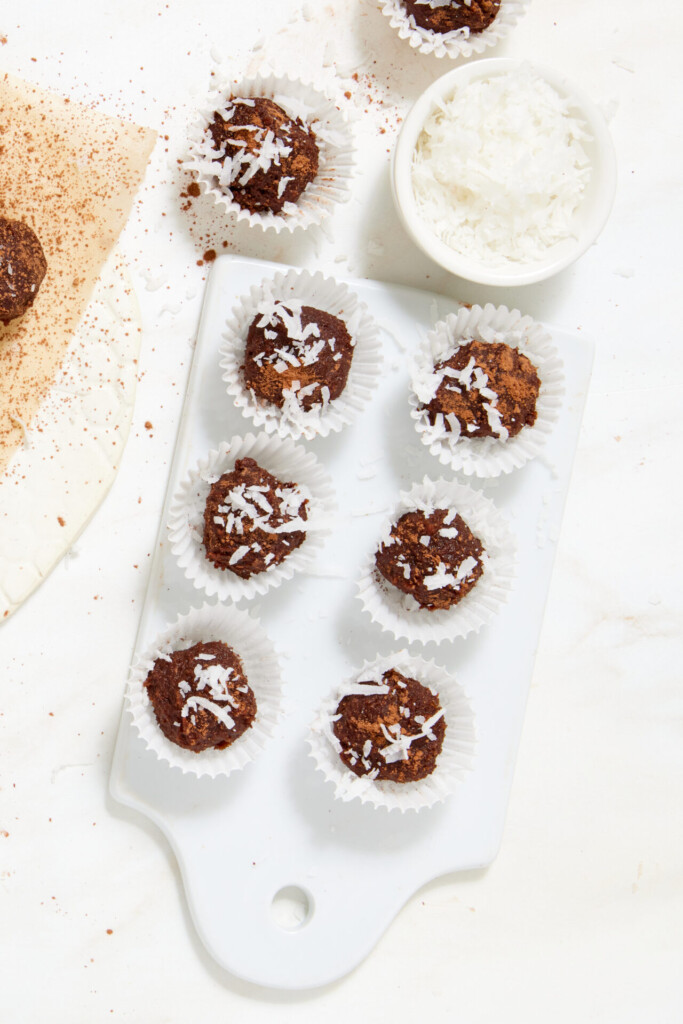 Chewy chocolate caramel candies made with dates, topped with shredded coconut in paper liners on a serving board, with a small bowl of coconut nearby.