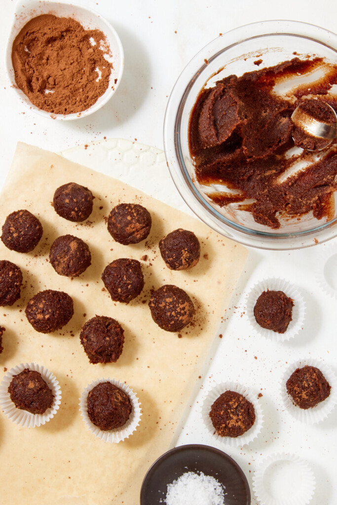 Chewy chocolate caramel candies made with dates in various stages of preparation, with some placed in paper liners, a bowl of cocoa powder, and a glass bowl of chocolate mixture.