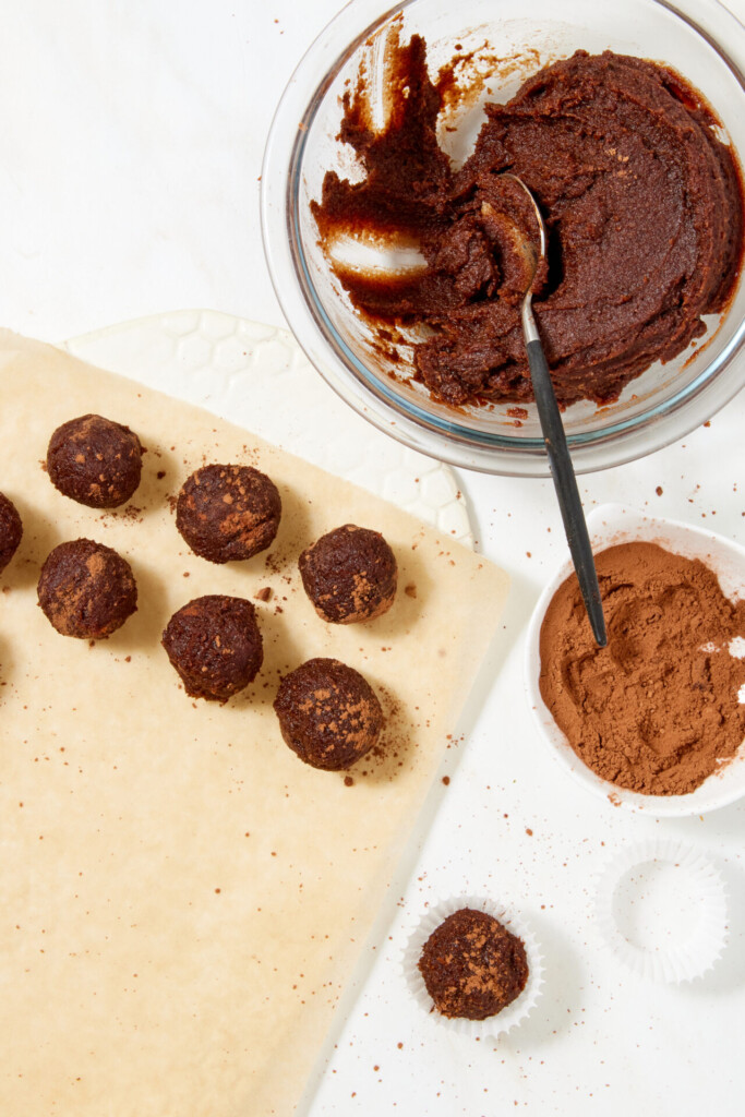 Several chewy chocolate caramel candies on parchment paper next to a mixing bowl of chocolate dough, a small bowl of cocoa powder, and a single truffle in a paper liner.