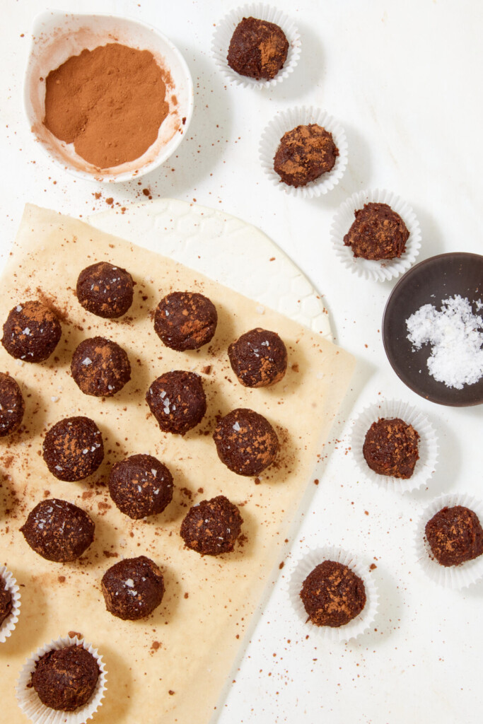 Top-down view of chewy chocolate caramel candies dusted with cocoa powder and sea salt, arranged on parchment paper and in small paper liners, with bowls of cocoa powder and sea salt nearby.