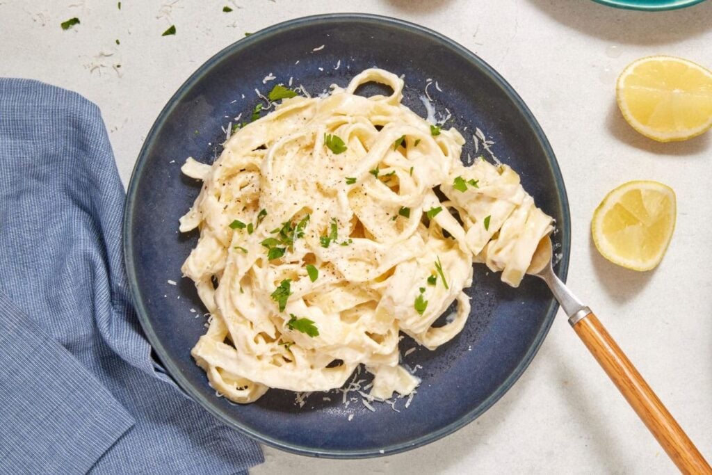 A plate of high-protein Cottage Cheese Fettuccine Alfredo garnished with parsley sits next to a blue napkin, lemon halves, a block of parmesan on a green plate, and a skillet of pasta on a light surface.