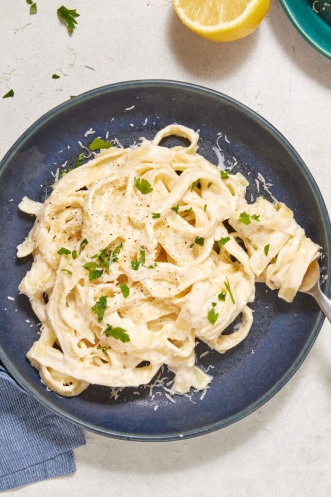 A plate of high-protein Cottage Cheese Fettuccine Alfredo garnished with parsley sits next to a blue napkin, lemon halves, a block of parmesan on a green plate, and a skillet of pasta on a light surface.